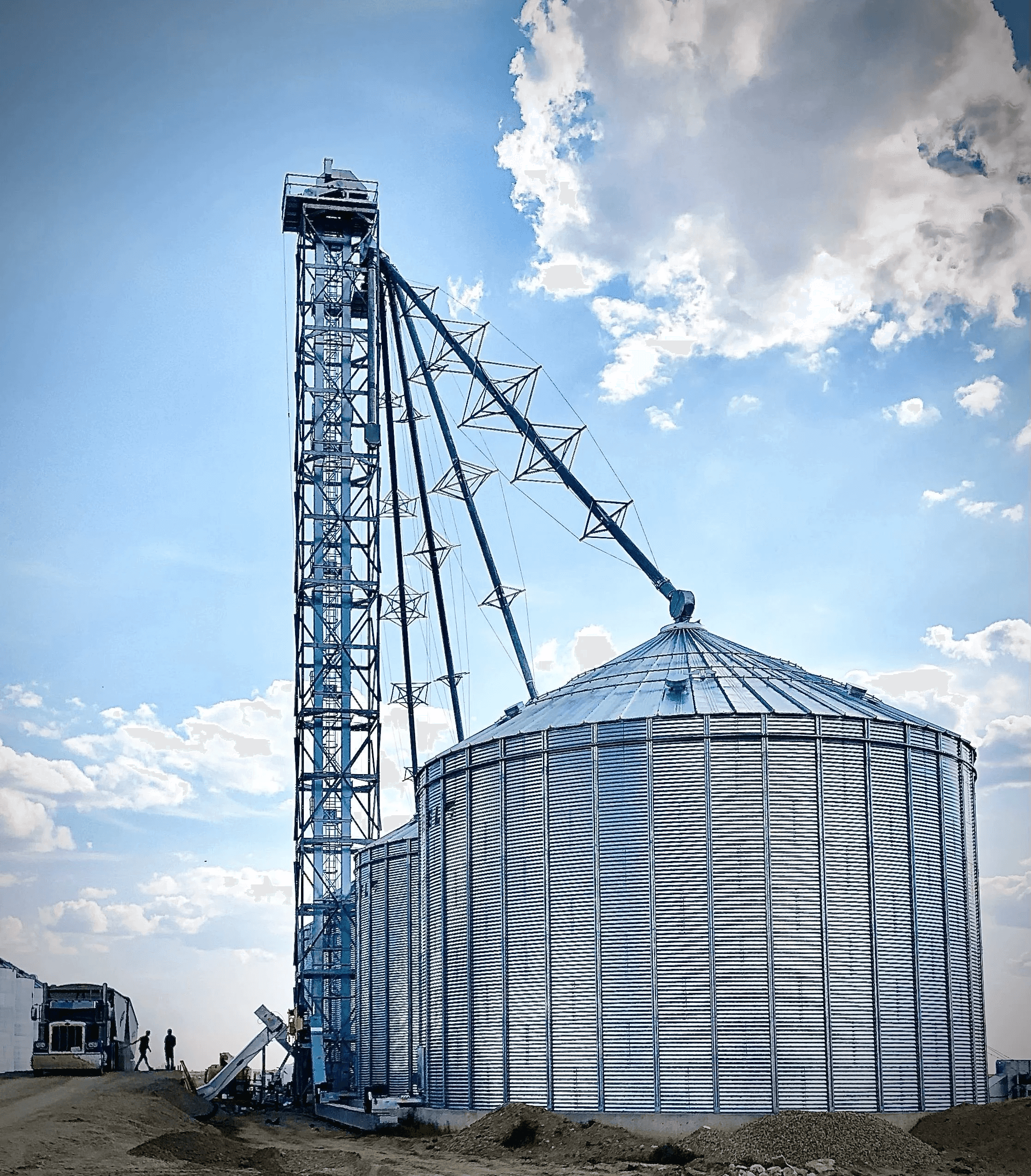 Galvanized bucket elevator support tower with spouting beside a large grain bin under a blue prairie sky