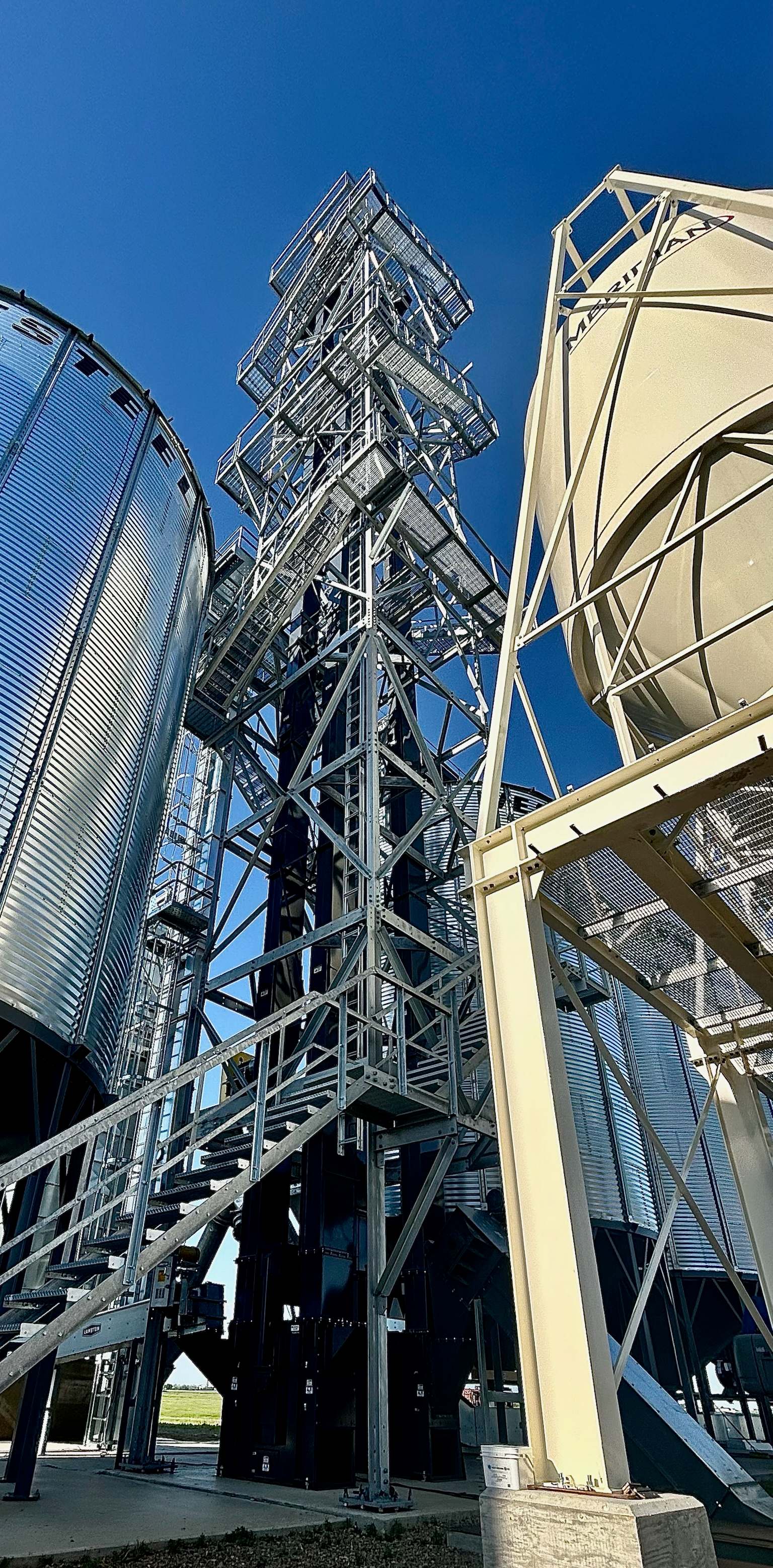Looking up a galvanized RapiTower with wrap-around stairs and landings between a grain bin and a hopper tank